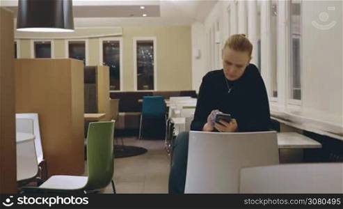 Cinemagraph - Young blond woman sitting alone at the table in empty cafe and browsing web on smart phone