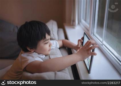 Cinematic portrait a happy young boy playing a game online on the tablet, Kid watching cartoon from internet, Child sitting on sofa next to window with a raining day in the morning