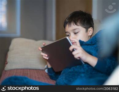 Cinematic portrait happy kid watching cartoon on tablet, Candid shot cute boy playing game online with friends on internet, Cozy Child wearing fluffy pajamas sitting on sofa relaxing at home in winter