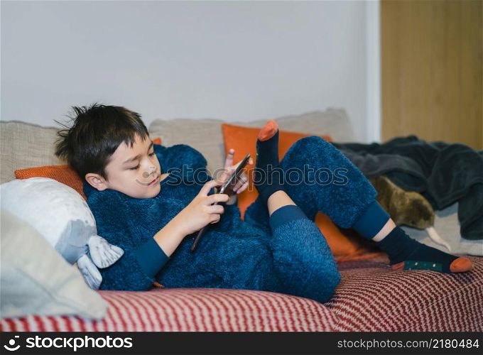 Cinematic portrait happy kid watching cartoon on tablet, Candid shot cute boy playing game online with friends on internet, Cozy Child wearing fluffy pajamas sitting on sofa relaxing at home