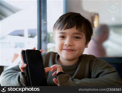 Cinematic portrait young boy playing game on mobile while waiting for food, Kid sitting in coffee shop sending text to friends, Child playing game online on phone.