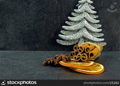 Cinnamon sticks, star anise, two slices dried orange and silver Christmas tree on a dark, stone table with shallow depth of field, as a Christmas decoration. Close up view from the side. Horizontal.