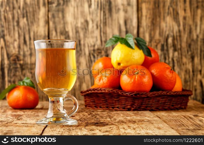 Citrus tea in a glass cup, warm healthy drink in the cool seasonon, background of mandarin and lemon fruits piled in a wicker basket on an old wooden table in blur, copy space.. Citrus tea in a glass cup on a background of mandarin and lemon fruits in blur.