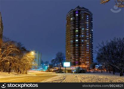 City landscape, high-rise residential apartment building against the background of the blue twilight of the winter sky and the city street of the evening park.. High-rise apartment building against the background of a city street in a winter city evening park covered with snow against a background of blue twilight.