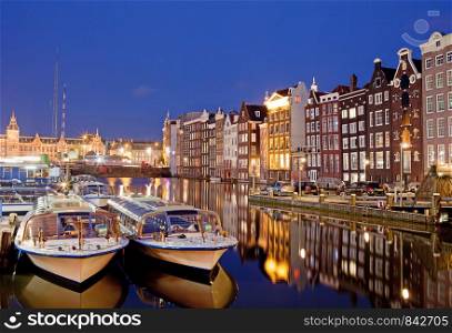 City of Amsterdam in Netherlands at night, historic apartment houses with reflections on water and boats ready for canal tours and cruises.