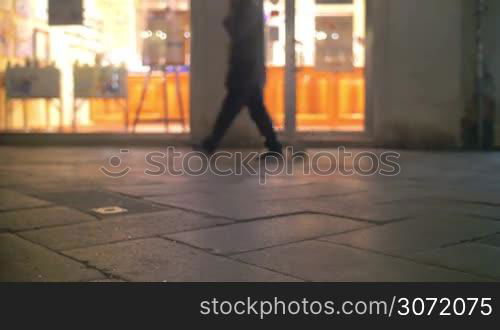 City people walking by the camera set in the street. There are human legs in shot, show window of a shop in the background.