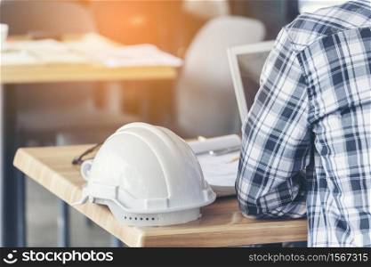 Civil construction engineer working with laptop at desk office with white yellow safety hard hat at office on construction site. Asian young man architecture project manager sitting at office on site