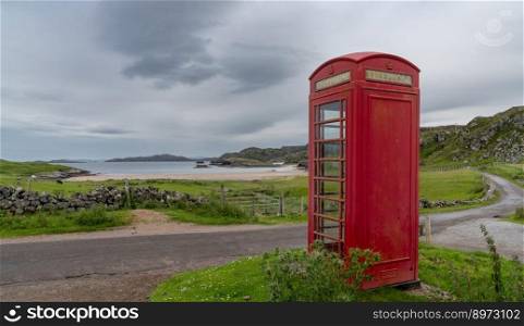 Clashnessie, United Kingdom - 28 June, 2022: typical red English phone booth in the middle of nowhere next to a picturesque beach in Scotland with a coastal road