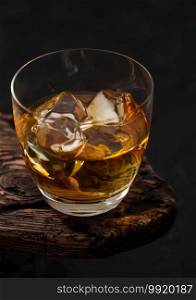 Classic glass of scotch malt whiskey with ice cubaes on top of wooden board and black background