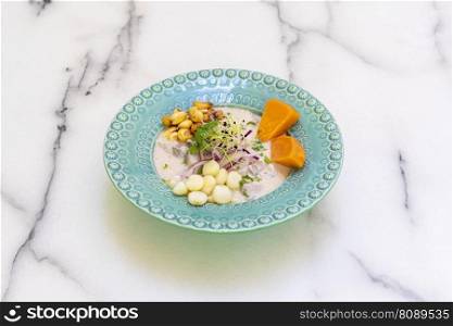 Classic Peruvian ceviche with marinated sea bass, grains of two varieties of corn, red onion, coriander, sweet potato and a beautiful blue plate on a white marble table