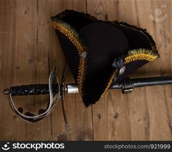 Classic pirate black felt captain's cocked hat and scabbard sword lying on a wooden background. Pirate Sword Hat