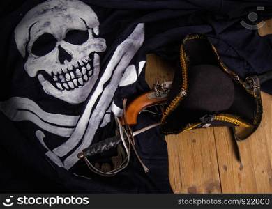 Classic pirate black felt captain's cocked hat with a pistol and a sword lying on a wooden floor next to the Jolly Roger flag. Pirate Sword Hat