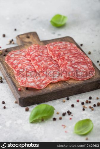 Classic salami slices with basil and pepper on wooden chopping board and light background.