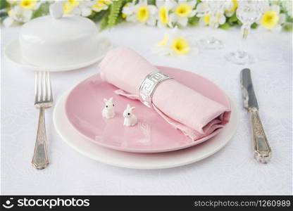 Classic serving for a Easter dinner with white and pink porcelain plates, pink napkin, silverware, two Easter rabbits and spring flowers are on a white tablecloth