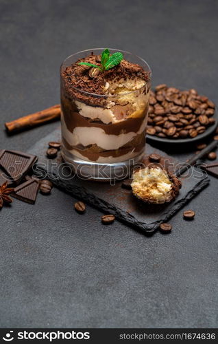 Classic tiramisu dessert in a glass cup and pieces of chocolate on stone cutting board on dark concrete background or table. Classic tiramisu dessert in a glass cup and pieces of chocolate on stone cutting board on dark concrete background