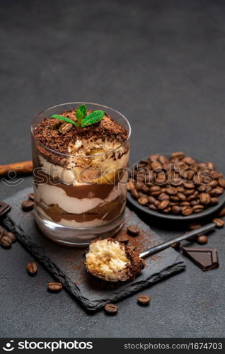 Classic tiramisu dessert in a glass cup and pieces of chocolate on stone cutting board on dark concrete background or table. Classic tiramisu dessert in a glass cup and pieces of chocolate on stone cutting board on dark concrete background