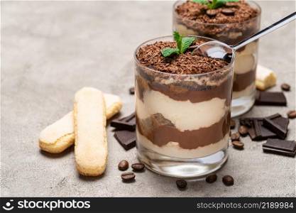 Classic tiramisu dessert in a glass cup, pieces of chocolate and savoiardi cookies on concrete background or table. Classic tiramisu dessert in a glass cup, pieces of chocolate and savoiardi cookies on concrete background