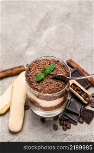Classic tiramisu dessert in a glass cup, pieces of chocolate and savoiardi cookies on concrete background or table. Classic tiramisu dessert in a glass cup, pieces of chocolate and savoiardi cookies on concrete background