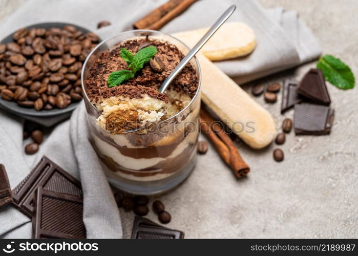 Classic tiramisu dessert in a glass cup, pieces of chocolate and savoiardi cookies on concrete background or table. Classic tiramisu dessert in a glass cup, pieces of chocolate and savoiardi cookies on concrete background