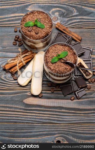 Classic tiramisu dessert in a glass cup, pieces of chocolate and savoiardi cookies on wooden background or table. Classic tiramisu dessert in a glass cup, pieces of chocolate and savoiardi cookies on wooden background