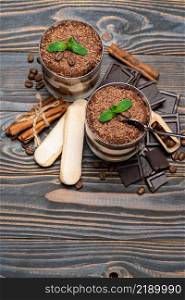 Classic tiramisu dessert in a glass cup, pieces of chocolate and savoiardi cookies on wooden background or table. Classic tiramisu dessert in a glass cup, pieces of chocolate and savoiardi cookies on wooden background