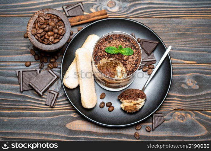 Classic tiramisu dessert in a glass cup, pieces of chocolate and savoiardi cookies on wooden background or table. Classic tiramisu dessert in a glass cup, pieces of chocolate and savoiardi cookies on wooden background