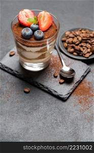 Classic tiramisu dessert with blueberries and strawberries in a glass on stone serving board on dark concrete background or table. Classic tiramisu dessert with blueberries and strawberries in a glass on stone serving board on dark concrete background