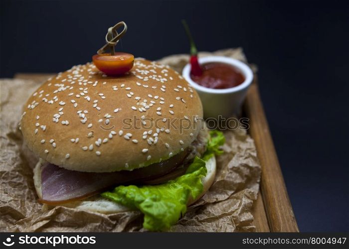 Classical American fresh juicy burger with chicken and ham on a wooden tray with a spicy chili sauce. Beautiful photo on a dark background. Classical American fresh juicy burger with chicken and ham on a wooden tray with a spicy chili sauce. Beautiful photo on a dark background.