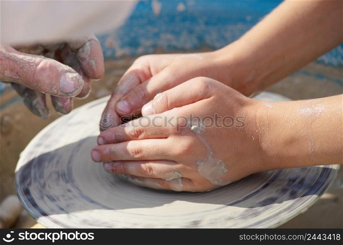 Clay stained children&rsquo;s hands work on a potter&rsquo;s wheel. Potter teaches a child to create a product from clay. Men&rsquo;s hands in the background. Clay stained children&rsquo;s hands work on a potter&rsquo;s wheel