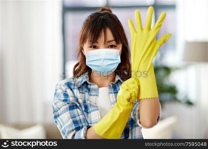 cleaning, health and hygiene concept - asian woman wearing protective medical mask for protection from virus disease and putting rubber gloves on at home. asian woman in protective mask and rubber gloves