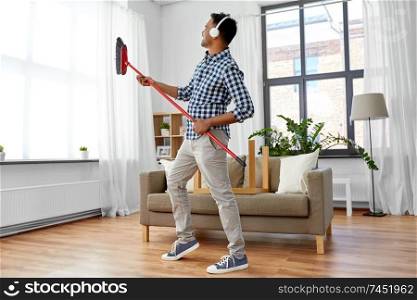 cleaning, housework and housekeeping concept - indian man in headphones with broom sweeping and having fun at home. man with broom cleaning and having fun at home