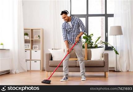 cleaning, housework and housekeeping concept - indian man in headphones with broom sweeping floor at home. man in headphones with broom cleaning at home