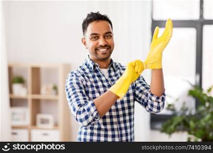 cleaning, housework and housekeeping concept - smiling indian man putting protective rubber gloves on at home. indian man putting protective rubber gloves on