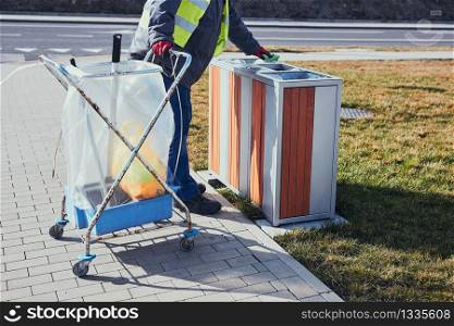 Cleaning service worker emptying the trash. Man wearing yellow vest, cleaning a bin, standing beside a cleaning cart. Real people, authentic situations