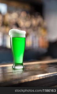 Clear glass of fresh natural green beer with extra foam on a wooden table on a background of pub. Happy St.Patrick &rsquo;s Day concept.. Glass of light green beer on a background of pub.