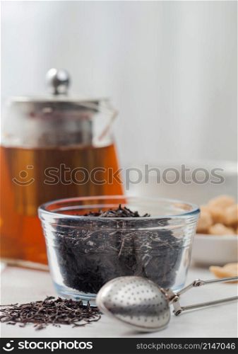 Clear glass teapot with loose organic black tea and cane sugar with white ceramic cup on light table background.