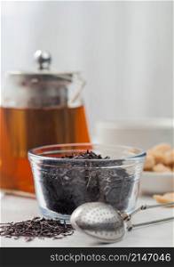 Clear glass teapot with loose organic black tea and cane sugar with white ceramic cup on light table background.