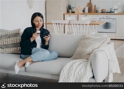 Client is purchasing online from home on quarantine. Young happy arab woman is resting on couch, holding smartphone and credit card. Girl is looking at screen and smiling. Money transfer app.. Client is purchasing online from home on quarantine. Young woman holding smartphone and credit card.