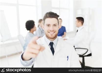 clinic, profession, gesture, people and medicine concept - happy male doctor pointing finger at you over group of medics meeting at hospital