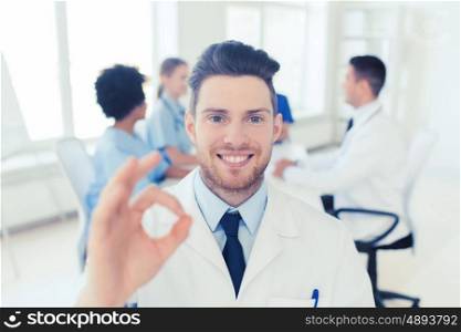 clinic, profession, gesture, people and medicine concept - happy male doctor showing ok hand sign over group of medics meeting at hospital