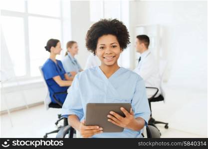 clinic, profession, people and medicine concept - happy african american female doctor or nurse with tablet pc computer over group of medics meeting at hospital