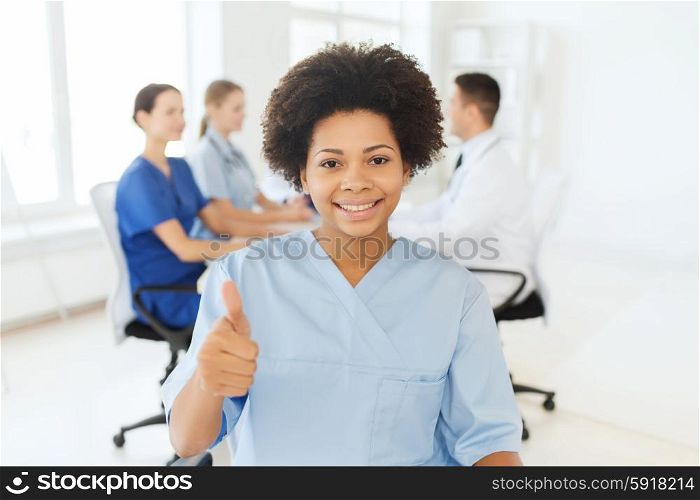 clinic, profession, people and medicine concept - happy african american female doctor or nurse over group of medics meeting at hospital showing thumbs up gesture