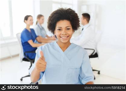 clinic, profession, people and medicine concept - happy african american female doctor or nurse over group of medics meeting at hospital showing thumbs up gesture