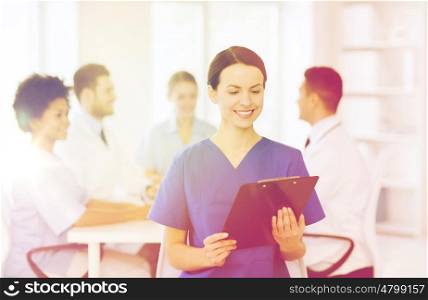 clinic, profession, people and medicine concept - happy female doctor or nurse with clipboard over group of medics meeting at hospital