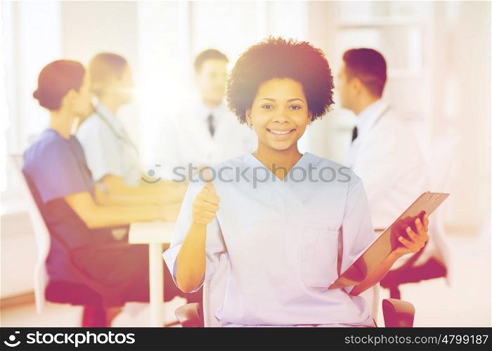 clinic, profession, people and medicine concept - happy female doctor or nurse with clipboard over group of medics meeting at hospital showing thumbs up gesture