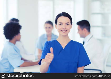 clinic, profession, people and medicine concept - happy female doctor or nurse over group of medics meeting at hospital showing thumbs up gesture