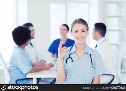 clinic, profession, people and medicine concept - happy female doctor over group of medics meeting at hospital showing ok hand sign. happy doctor over group of medics at hospital