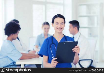 clinic, profession, people and medicine concept - happy female doctor with clipboard over group of medics meeting at hospital. happy doctor over group of medics at hospital