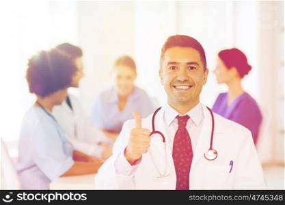 clinic, profession, people and medicine concept - happy male doctor over group of medics meeting at hospital showing thumbs up gesture