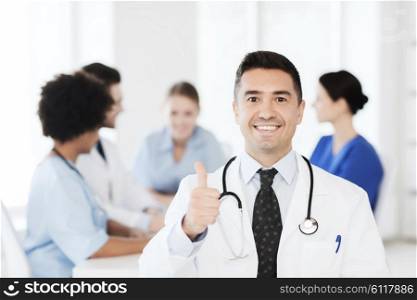 clinic, profession, people and medicine concept - happy male doctor over group of medics meeting at hospital showing thumbs up gesture
